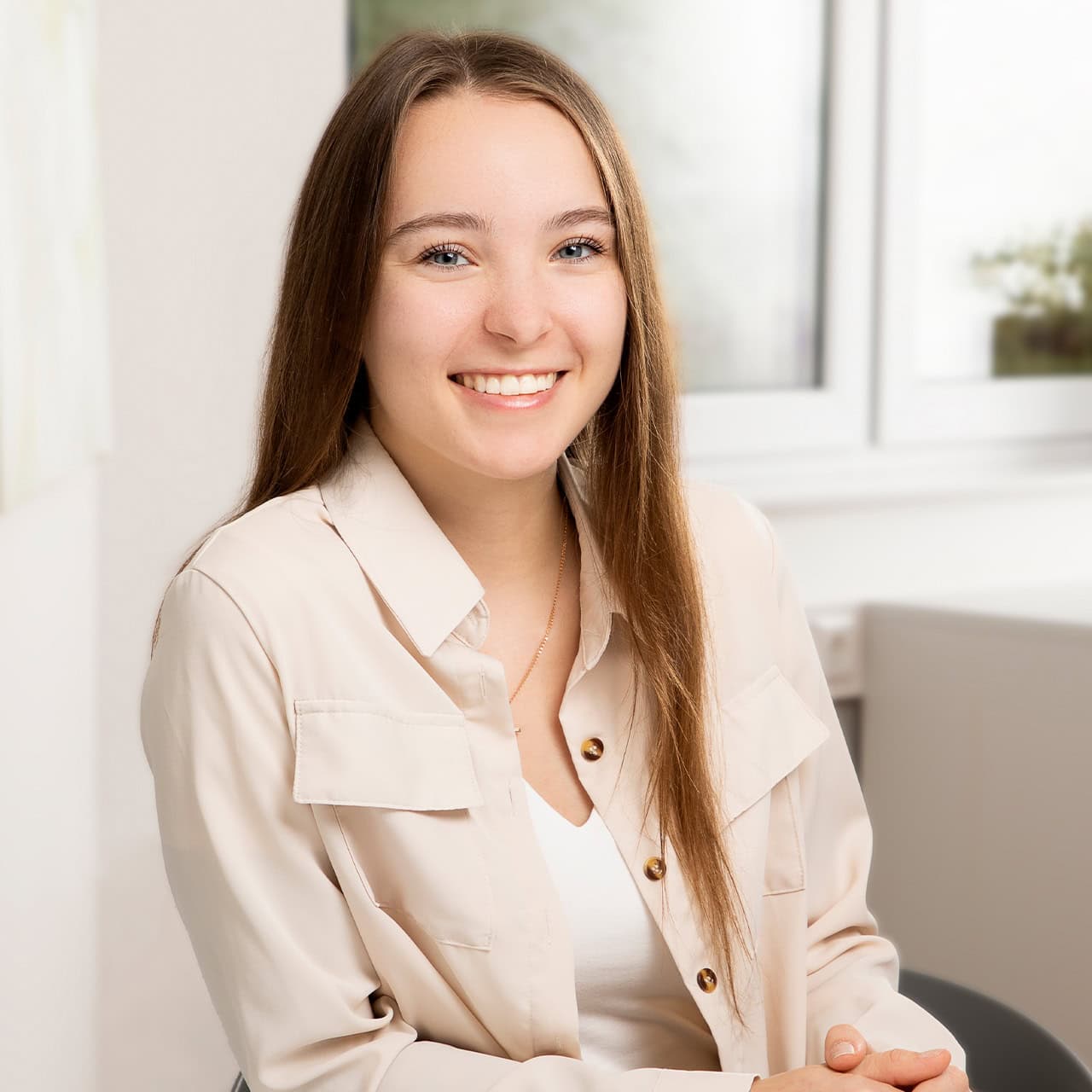Junge Frau lächelt freundlich, trägt cremefarbenes Hemd, weißes Top, lange braune Haare, sitzt.
