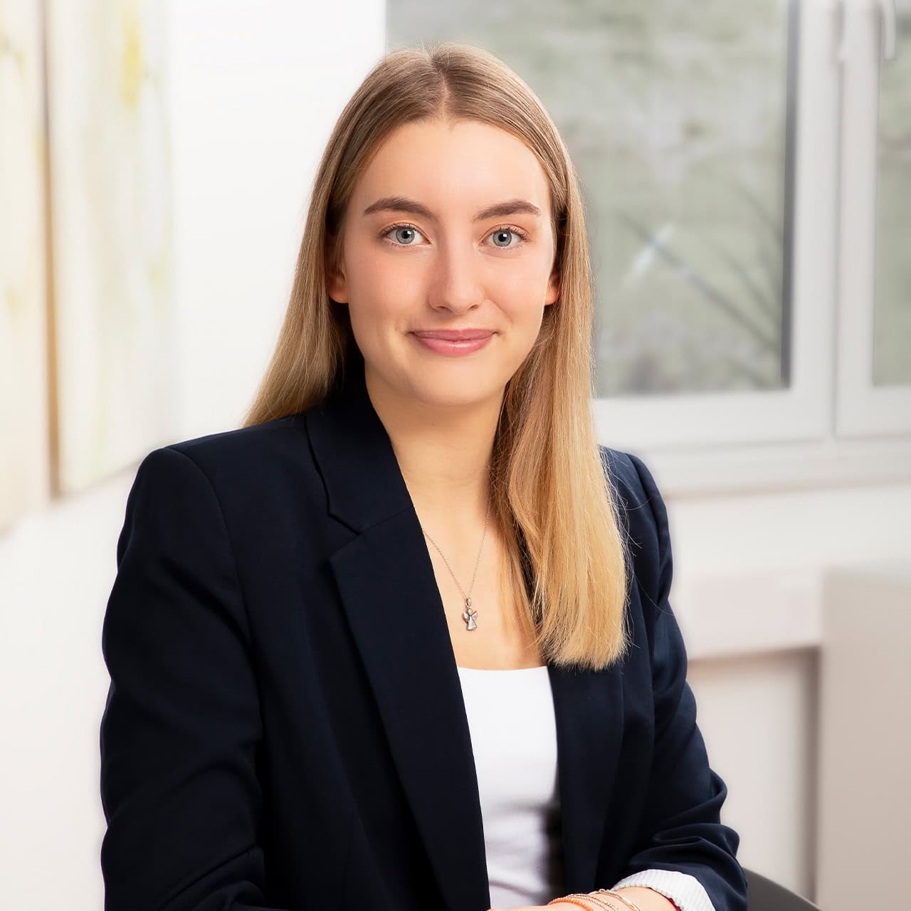 Eine lächelnde Frau mit blonden Haaren, in einem schwarzen Blazer vor einem Fenster sitzend.