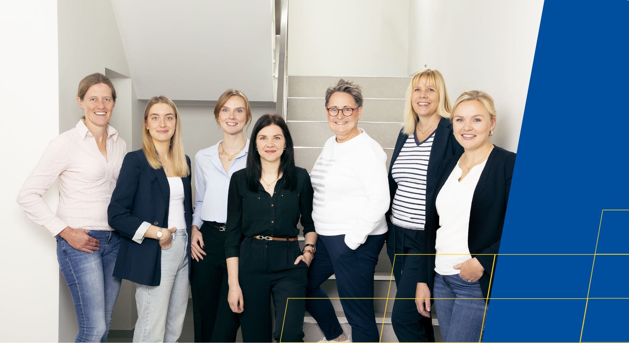 Eine Gruppe von sieben Frauen in lächelnder Pose vor einer Treppe, teils in Jeans und Blazern.