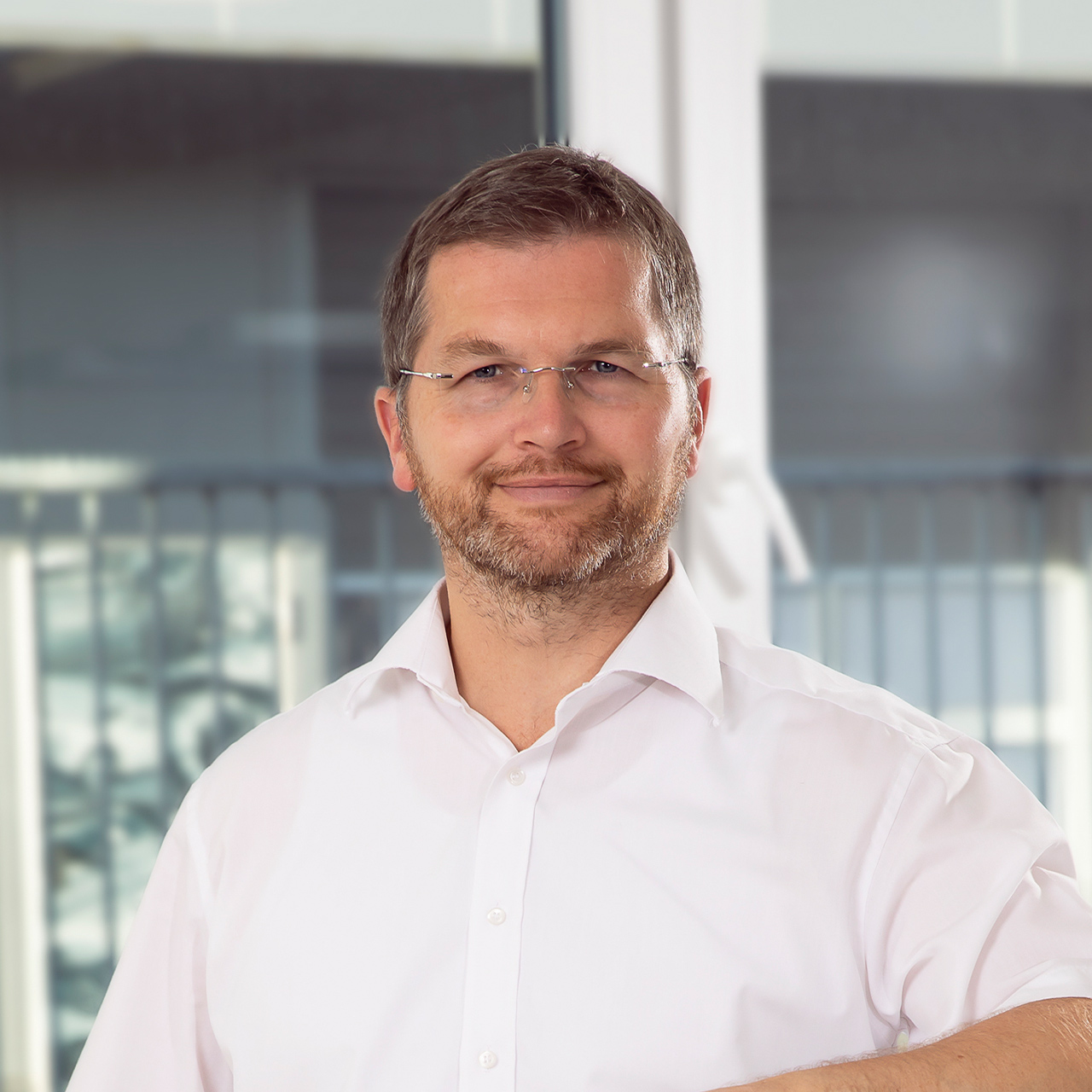 Eine lächelnde Person mit Brille und Bart trägt ein weißes Hemd in hellem Raum
